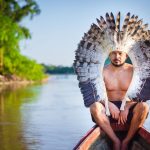 Homem indígena sentado em um barco no rio, usando um cocar de penas, em Rio Branco, no Brasil.