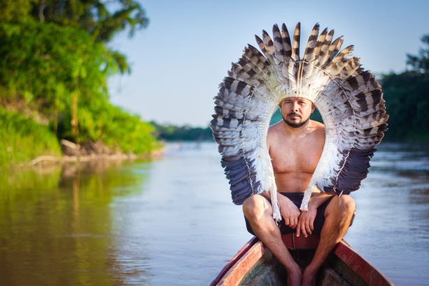 Homem indígena sentado em um barco no rio, usando um cocar de penas, em Rio Branco, no Brasil.