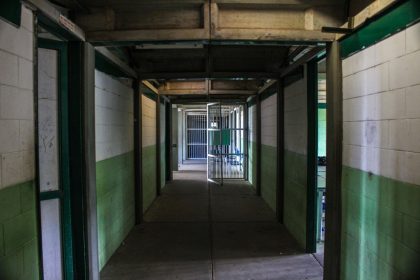 Corredor vazio de uma prisão, com portas de celas abertas e iluminação institucional.