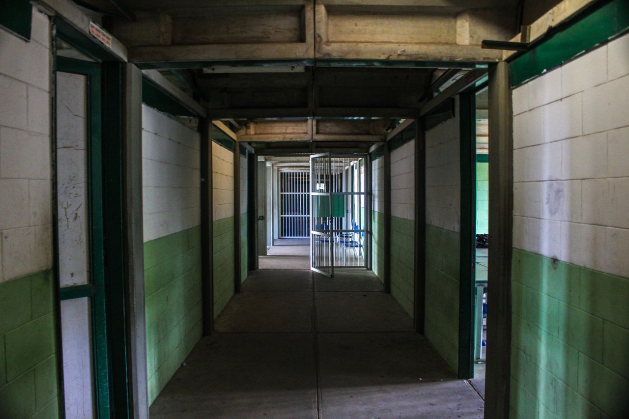 Corredor vazio de uma prisão, com portas de celas abertas e iluminação institucional.