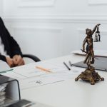 Mesa de trabalho jurídico com estátua da Justiça, documentos e um laptop.